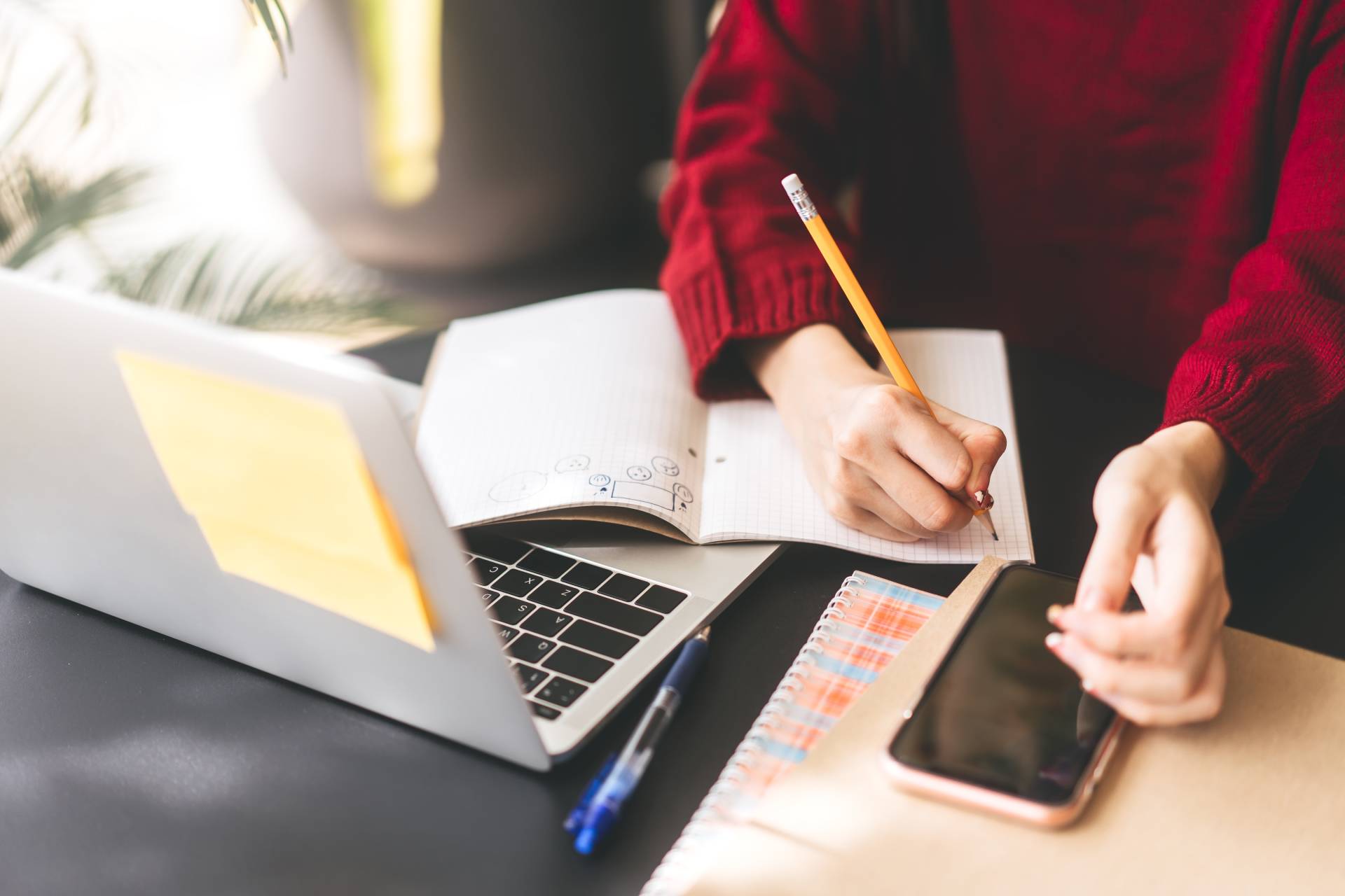 Photograph of a student making notes in an exercise book with a laptop and a phone