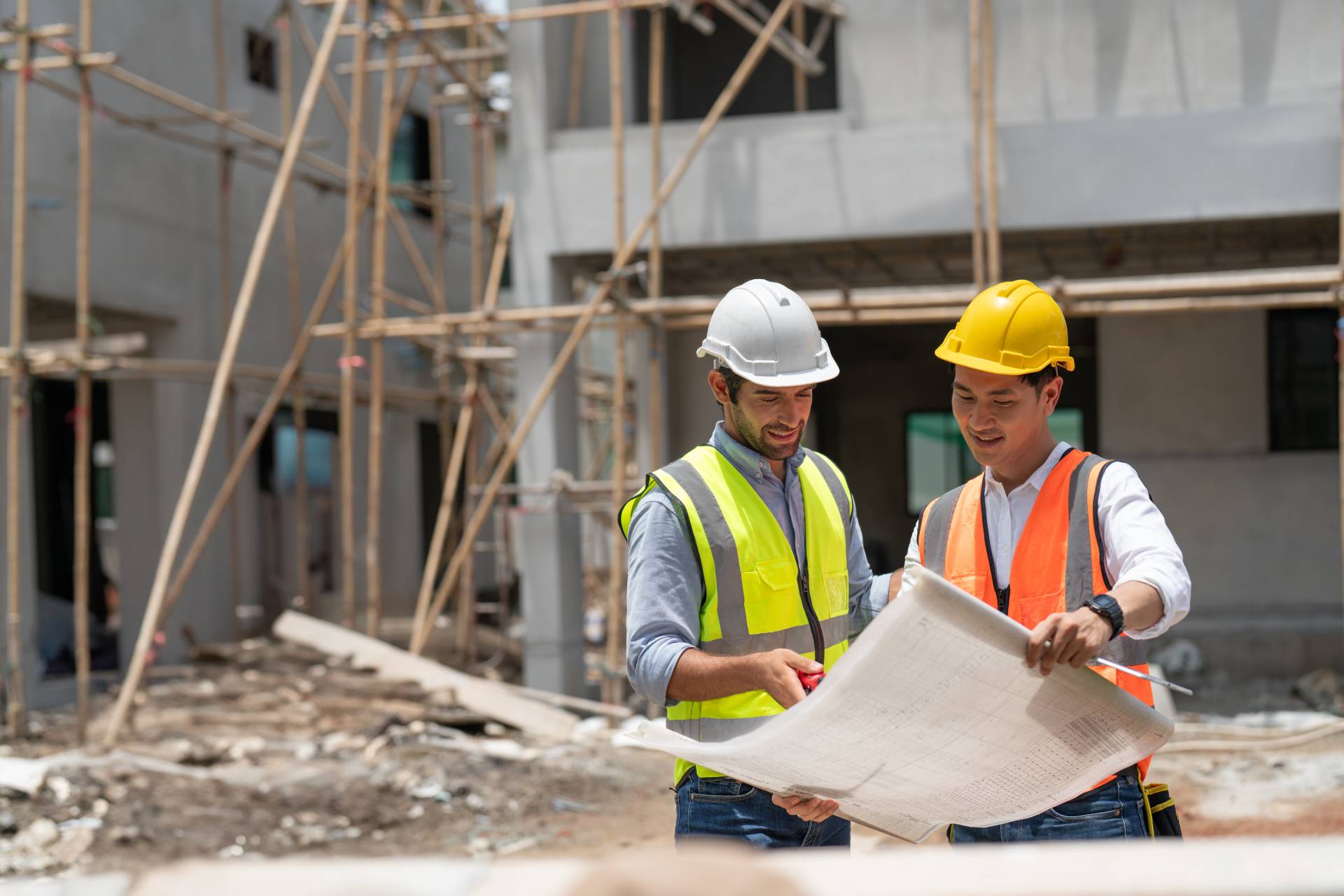 Photograph of two men in high-vis vests and safety helmets looking at blueprints on a construction site