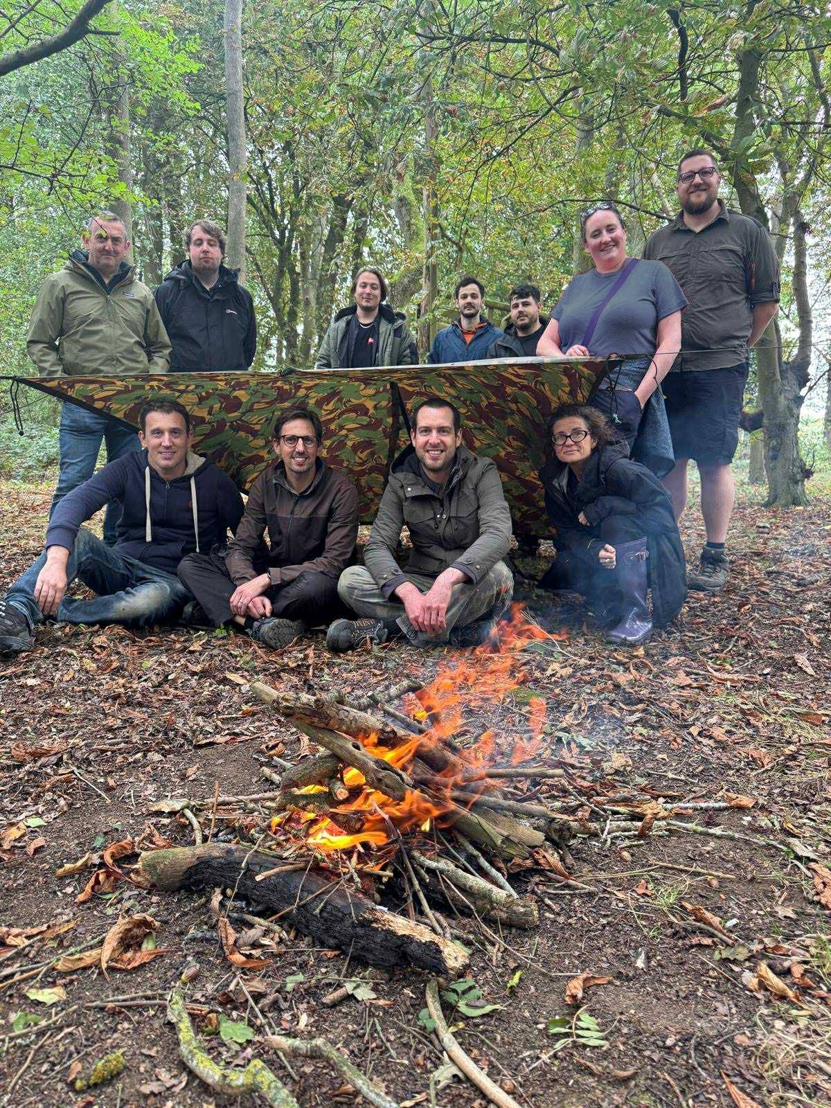 Photograph of the Hero team at a team building day in a forest, eleven people around a shelter they've built, in front of a campfire